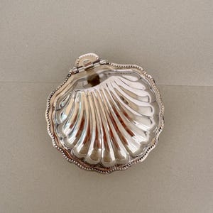 May include: A silver-toned metal clam shell dish with a hinged lid. The exterior surface features a polished, reflective finish with deep, radiating grooves and a beaded edge detail along the perimeter. The object is shaped like a scallop shell.