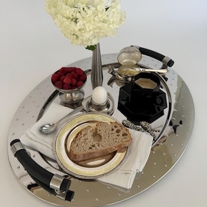 May include: An oval silver-toned serving tray with black handles and a dotted pattern, holding a white hydrangea bouquet, a bowl of raspberries, a boiled egg in a cup, a slice of bread on a plate, a black cup, and a silver spoon on a napkin.