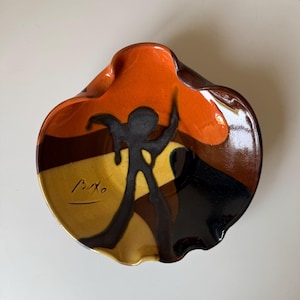 Puede incluir: Un plato de cerámica con un diseño abstracto único. El plato presenta una paleta de colores naranja, marrón, negro y amarillo. Una silueta negra de una figura está pintada en el plato. El plato está firmado con el nombre "Buxo".