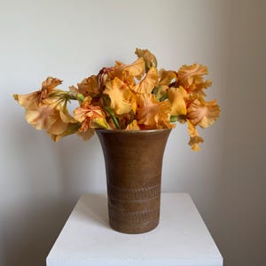 May include: A brown vase with a textured surface holds a bouquet of orange irises. The vase has a flared top and decorative horizontal bands. The flowers are in full bloom, with delicate petals and a warm color palette.