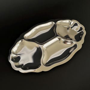 May include: A polished, silver-coloured serving tray with three sections. The tray has an unusual, organic form with softly curved edges. The reflective surface creates highlights and shadows, enhancing its visual appeal. Ideal for serving snacks or appetizers.