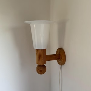 Może przedstawiać: Lampa ścienna z białym, szklanym kloszem w kształcie tulipana. Lampa posiada drewnianą podstawę i ramię, z okrągłym drewnianym akcentem. Widoczny jest przewód zasilający z włącznikiem/wyłącznikiem.