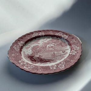 May include: Oval serving platter with a pink and white floral border and a central landscape scene. The platter has a scalloped edge and two small handles. The design features figures and buildings in a pastoral setting.