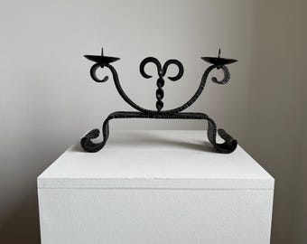 May include: Black wrought iron candle holder with two candle cups. The decorative piece features scrolled and looped designs, with a central flourish. The base has curved ends, and the piece is displayed on a white pedestal.