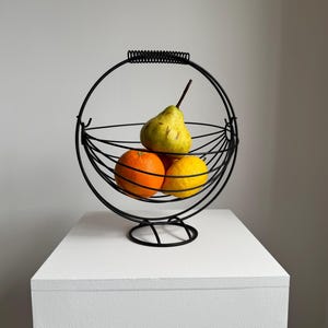 May include: A black wire fruit basket with a circular design, holding a green pear, an orange, and a yellow lemon. The basket sits on a white surface. The basket has a handle at the top and a round base.