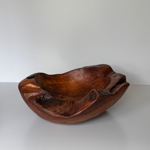 Può includere: Una ciotola in legno fatta a mano con una forma naturale e organica. La ciotola è realizzata in legno marrone scuro, che mostra la venatura e la consistenza. I bordi sono irregolari, aggiungendo fascino rustico. Questa ciotola decorativa è adatta a vari usi.