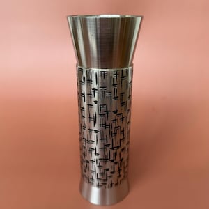 May include: A tall, cylindrical metal vase with a brushed finish. The body features a black, geometric pattern. The top flares out, and the base is a smaller, matching metal. The background is a solid peach color.