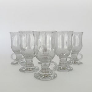 Può includere: Un set di cinque tazze in vetro trasparente, ciascuna dotata di base a piedistallo, stelo corto e manico curvo. Le tazze presentano una forma a coppa liscia e arrotondata per bevande calde, disposte in gruppo su uno sfondo neutro.