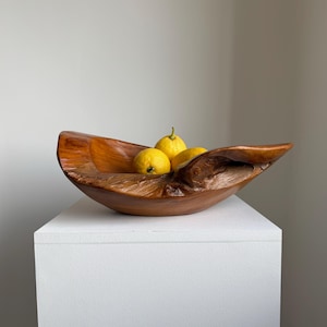 Può includere: Una ciotola di legno fatta a mano con una forma naturale e organica. Contiene tre limoni gialli. Il legno ha un colore marrone caldo e ricco, con venature visibili. La ciotola è esposta su un piedistallo bianco. Circa 30 cm di lunghezza.