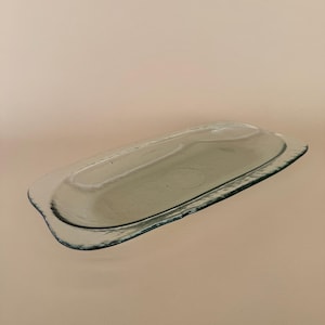 May include: A clear, rectangular glass tray with rounded edges. The tray has a slightly raised, oval-shaped interior. The glass has a subtle green tint, and the edges are slightly textured.