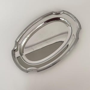 Op de afbeelding: Een ovaal zilverkleurig metalen dienblad met een gepolijst, reflecterend oppervlak. Het dienblad heeft een verhoogde, geschulpte rand met een decoratief patroon van kleine, gegraveerde kruisvormige markeringen langs de rand.
