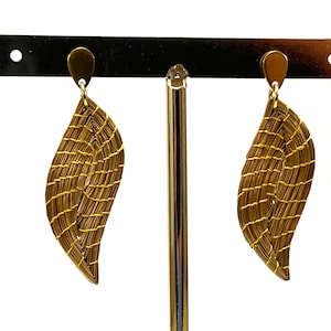 Peut inclure: Une paire de boucles d'oreilles en forme de feuille dorées avec un motif tissé. Les boucles d'oreilles sont suspendues à un présentoir noir.