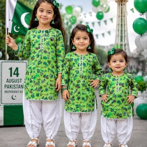 May include: Three children wearing matching green patterned long-sleeved tunics with white trousers and green hair accessories. The tunics feature a repeating graphic print, and the white trousers have a gathered, loose-fitting design.