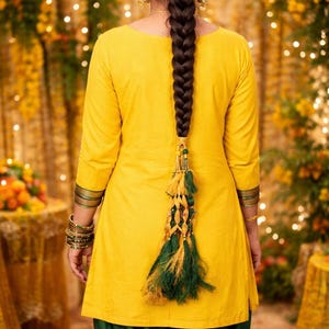 May include: A woman wearing a yellow tunic and green pants. Her long, dark hair is styled in a braid, adorned with a decorative tassel. The outfit includes gold bangles and trim, creating a vibrant and festive look.
