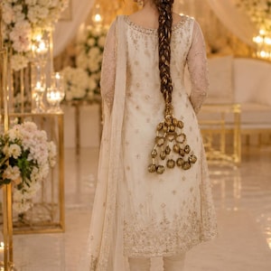 May include: An off-white long-sleeved tunic with intricate gold embroidery and beadwork, paired with matching fitted trousers and a sheer dupatta. The garment features a scoop neckline and a long, braided hairstyle adorned with gold ornaments.