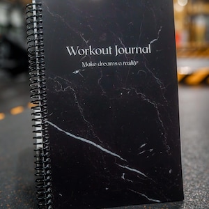Pode incluir: Um diário de treino com padrão de mármore preto e branco com encadernação em espiral. A capa apresenta o texto "Workout Journal" acima da frase "Make dreams a reality" em uma fonte branca e elegante. O diário está aberto.