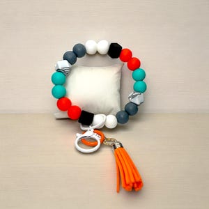 May include: A colorful beaded bracelet with a tassel. The bracelet features a variety of beads in teal, red, gray, white, and black. The bracelet has a white clasp and an orange tassel. The bracelet is approximately 8 inches in circumference.