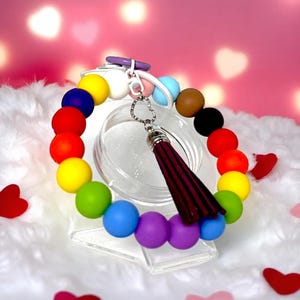 May include: A rainbow-colored beaded keychain bracelet with a burgundy tassel. The bracelet features a variety of vibrant sphere-shaped beads in red, orange, yellow, green, blue, and purple. The keychain has a silver-colored clasp.