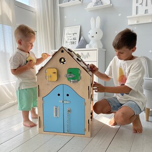 Puede incluir: Dos ni&ntilde;os juegan con una casa de juguete de madera con una puerta azul y varios pestillos y cerraduras. La casa tiene una campana en el techo y una ventana amarilla y verde. La casa est&aacute; sobre un suelo de madera claro.