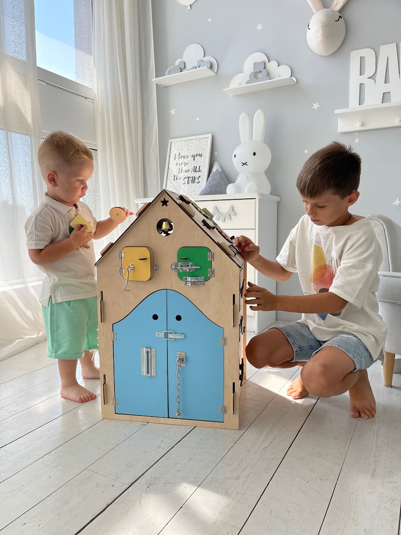 Puede incluir: Dos ni&ntilde;os juegan con una casa de juguete de madera con una puerta azul y varios pestillos y cerraduras. La casa tiene una campana en el techo y una ventana amarilla y verde. La casa est&aacute; sobre un suelo de madera claro.