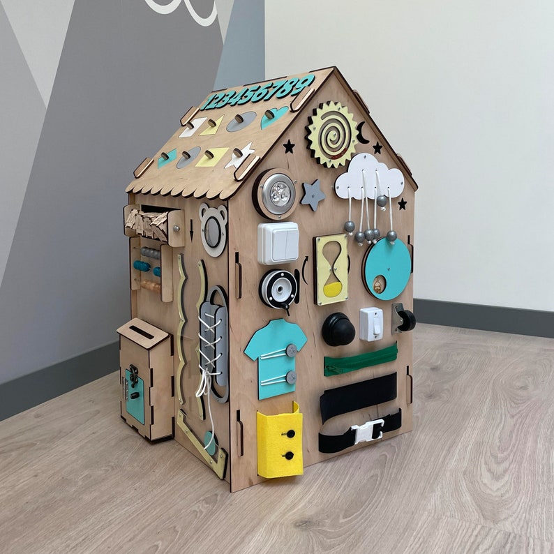 Puede incluir: Un tablero de actividades de madera con forma de casa con varias texturas y actividades para ni&ntilde;os. Cuenta con un interruptor de luz, una puerta con pestillo, una cremallera, una hebilla, un engranaje, una nube con cuentas colgantes y una secuencia de n&uacute;meros del 1 al 9.
