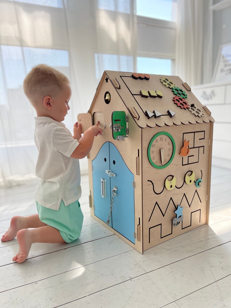 Puede incluir: Una casa de juegos de madera con una puerta azul, una esfera de reloj y varias otras actividades para que los ni&ntilde;os interact&uacute;en. La casa de juegos est&aacute; hecha de madera natural y tiene un acabado marr&oacute;n claro.