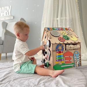 Puede incluir: Un tablero de actividades de madera con forma de casa con diversas actividades para ni&ntilde;os peque&ntilde;os, que incluyen un reloj, engranajes, un xil&oacute;fono, una cremallera, un zapato con cordones y un cocodrilo. Un ni&ntilde;o peque&ntilde;o est&aacute; jugando con el tablero de actividades.
