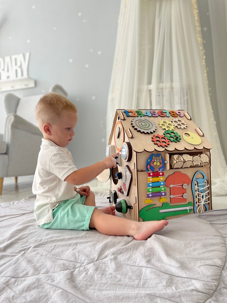 Puede incluir: Un tablero de actividades de madera con forma de casa con diversas actividades para ni&ntilde;os peque&ntilde;os, que incluyen un reloj, engranajes, un xil&oacute;fono, una cremallera, un zapato con cordones y un cocodrilo. Un ni&ntilde;o peque&ntilde;o est&aacute; jugando con el tablero de actividades.