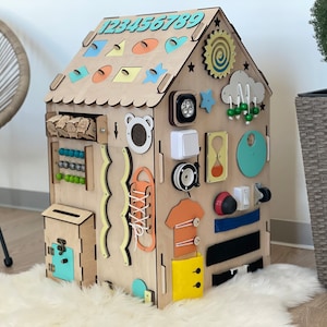 Puede incluir: Un tablero de actividades de madera con forma de casa con diversas actividades para ni&ntilde;os, que incluyen una secuencia de n&uacute;meros, un &aacute;baco, un cord&oacute;n, un buz&oacute;n, un interruptor de luz, una cremallera y un sol con un dise&ntilde;o en espiral.