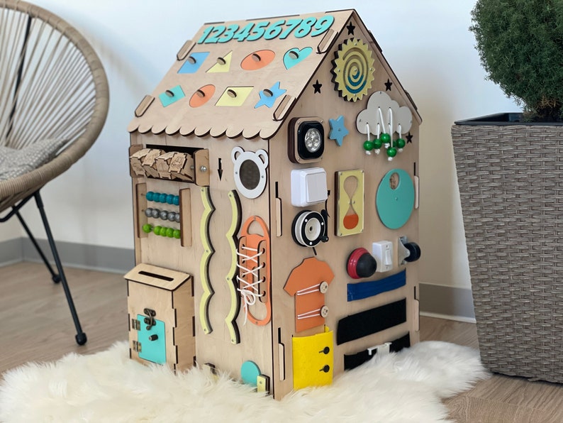 Puede incluir: Un tablero de actividades de madera con forma de casa con diversas actividades para ni&ntilde;os, que incluyen una secuencia de n&uacute;meros, un &aacute;baco, un cord&oacute;n, un buz&oacute;n, un interruptor de luz, una cremallera y un sol con un dise&ntilde;o en espiral.