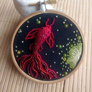 Peut inclure: Art de broderie en cercle représentant un poisson rouge avec une queue perlée sur un fond noir avec des accents verts et blancs.