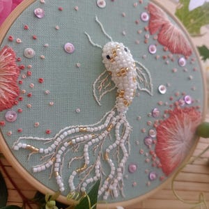 Peut inclure: Un poisson koï blanc et doré brodé sur un tissu bleu clair avec des paillettes roses et blanches. La broderie est encadrée dans un cercle en bois.