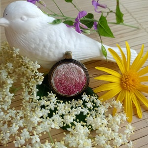 Peut inclure: Un pendentif brodé rose et blanc avec un cadre en bronze, entouré de fleurs blanches et d'une fleur jaune.