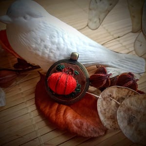 Peut inclure: Un pendentif rond en métal brun avec un motif de citrouille brodé en rouge. Le pendentif est sur une surface en bois brun tressé avec des feuilles séchées et une figurine d'oiseau blanche.