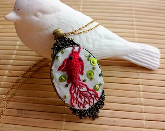 Può includere: Una collana con un ciondolo a forma di pesce rosso ricamato di colore rosso con accenti verdi e bianchi. Il ciondolo è in una cornice ovale in bronzo con una catena d'oro.