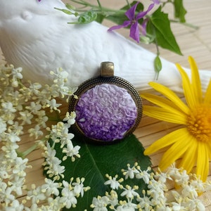 Peut inclure: Un pendentif en forme de fleur brodée de couleur violet et blanc avec un cadre en bronze. Le pendentif est entouré de fleurs blanches et de feuilles vertes.