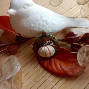 Peut inclure: Un pendentif artisanal avec un motif de citrouille brodée. Le pendentif est serti dans un cadre de couleur bronze et repose sur des feuilles d'automne. Une figurine d'oiseau en céramique blanche est en arrière-plan, suggérant un thème d'automne ou de récolte.