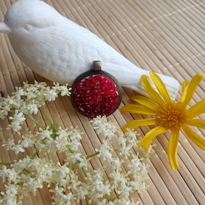 Peut inclure: Un pendentif brodé rouge avec un cadre en bronze, entouré de fleurs blanches et d'une fleur jaune sur un tapis en bambou.