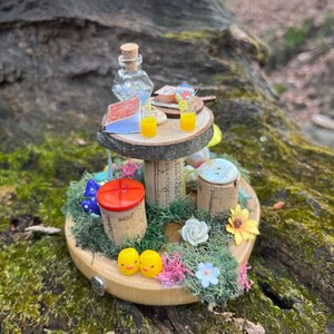 Könnte beinhalten: Eine Miniatur-Feengarten-Szene mit einem Holztisch, zwei Gläsern Orangensaft, einem Buch, einer Flasche und einem Korkuntergrund mit Moos, Blumen und zwei gelben Küken.