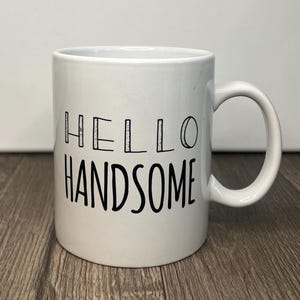 May include: White ceramic mug with a curved handle. The mug features the words "HELLO HANDSOME" in black, bold lettering. The mug is sitting on a wooden surface.