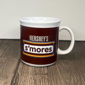 Może przedstawiać: Kubek z białym uchwytem, ciemnobrązowym zewnętrzem i białym obramowaniem. Kubek posiada logo Hershey's s'mores w kolorze białym i niebieskim. Kubek przeznaczony jest do gorących napojów.