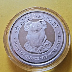 May include: A circular silver coin encased in a clear plastic protective capsule. The coin features a koala in the center with the text 5th ANNIVERSARY and ONE TROY OUNCE .999 FINE SILVER inscribed along the outer edge of the silver surface.