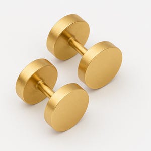 May include: Two gold-colored dumbbell-shaped cabinet knobs. Each knob has a cylindrical connecting bar between two flat, circular discs. The knobs are made of a brushed metal material and are set against a white background.