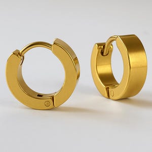 Può includere: Un paio di orecchini a cerchio color oro. Gli orecchini hanno un design spesso e arrotondato e una chiusura a cerniera. Il metallo ha una finitura lucida e lucidata. Gli orecchini sono esposti su uno sfondo bianco.