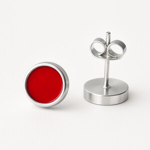 May include: A pair of stud earrings. Each earring has a bright red circular centre set in a silver-coloured metal frame. One earring is in focus, while the other shows the post and backing.