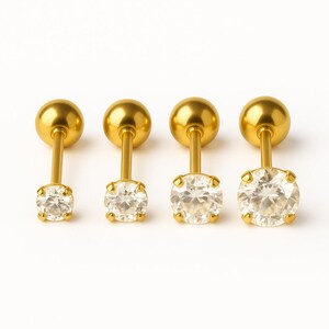 May include: Four gold-toned stud earrings with clear round gemstones. The earrings are arranged in a row from left to right.