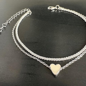 Może przedstawiać: Srebrna bransoletka na kostkę z podwójną warstwą. Górna warstwa ma teksturowany, błyszczący wygląd, a dolna warstwa zawiera łańcuszek z zawieszką w kształcie serca. Bransoletka na kostkę ma regulowany łańcuszek.