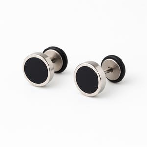 Può includere: Due orecchini a bottone in acciaio inossidabile con centri circolari neri. Gli orecchini hanno un perno e un retro in gomma nera. Presentati su sfondo bianco.