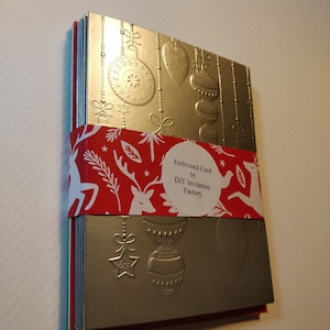 May include: A stack of silver and red embossed Christmas cards featuring a reindeer and ornament design. The cards are wrapped with red and white patterned paper and a white circular label that reads "Embossed Card by DIY Invitation Factory".