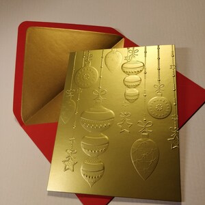 May include: A gold Christmas card with a red envelope. The card features a design of embossed gold baubles hanging from a string of beads.